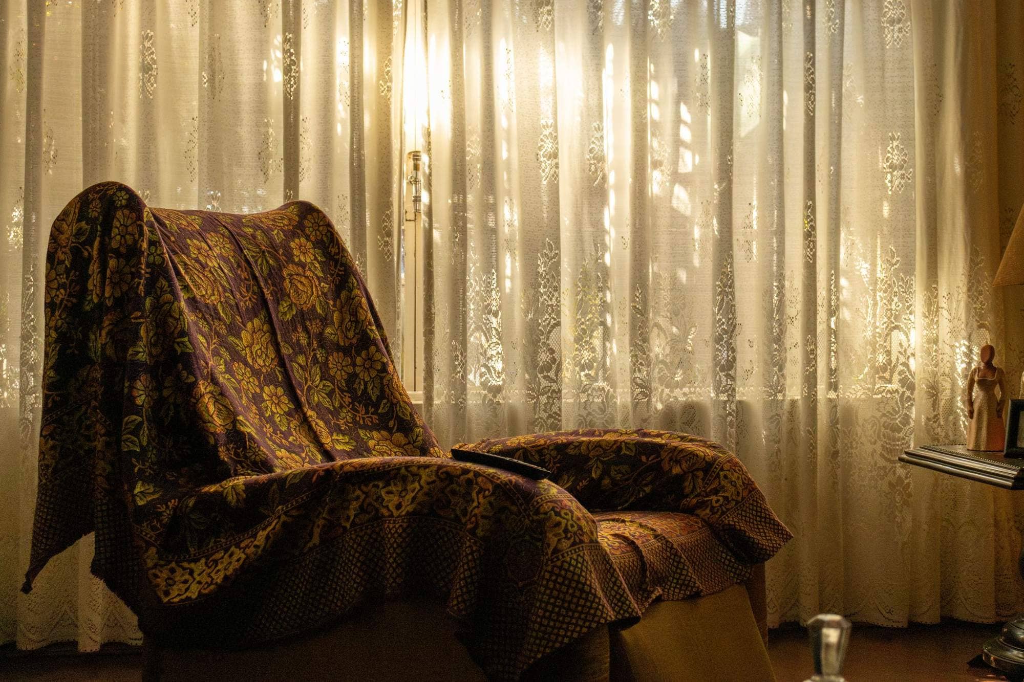 An armchair with a floral throw beside a lace-curtained window in low afternoon sun An armchair with a floral throw beside a lace-curtained window in low afternoon sun